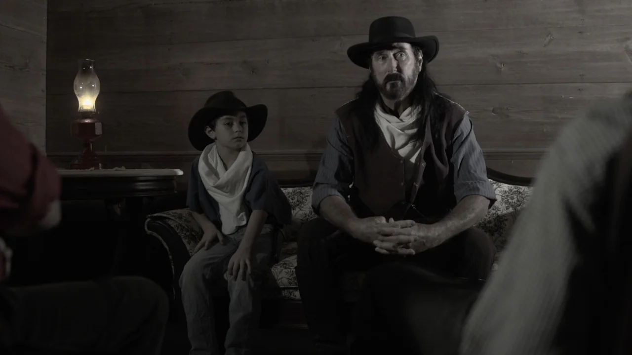 The Dalton Gang (2020) movie still: medium — A rustic wooden room contains a vintage patterned sofa and an oil lamp. A man and a young boy sit…