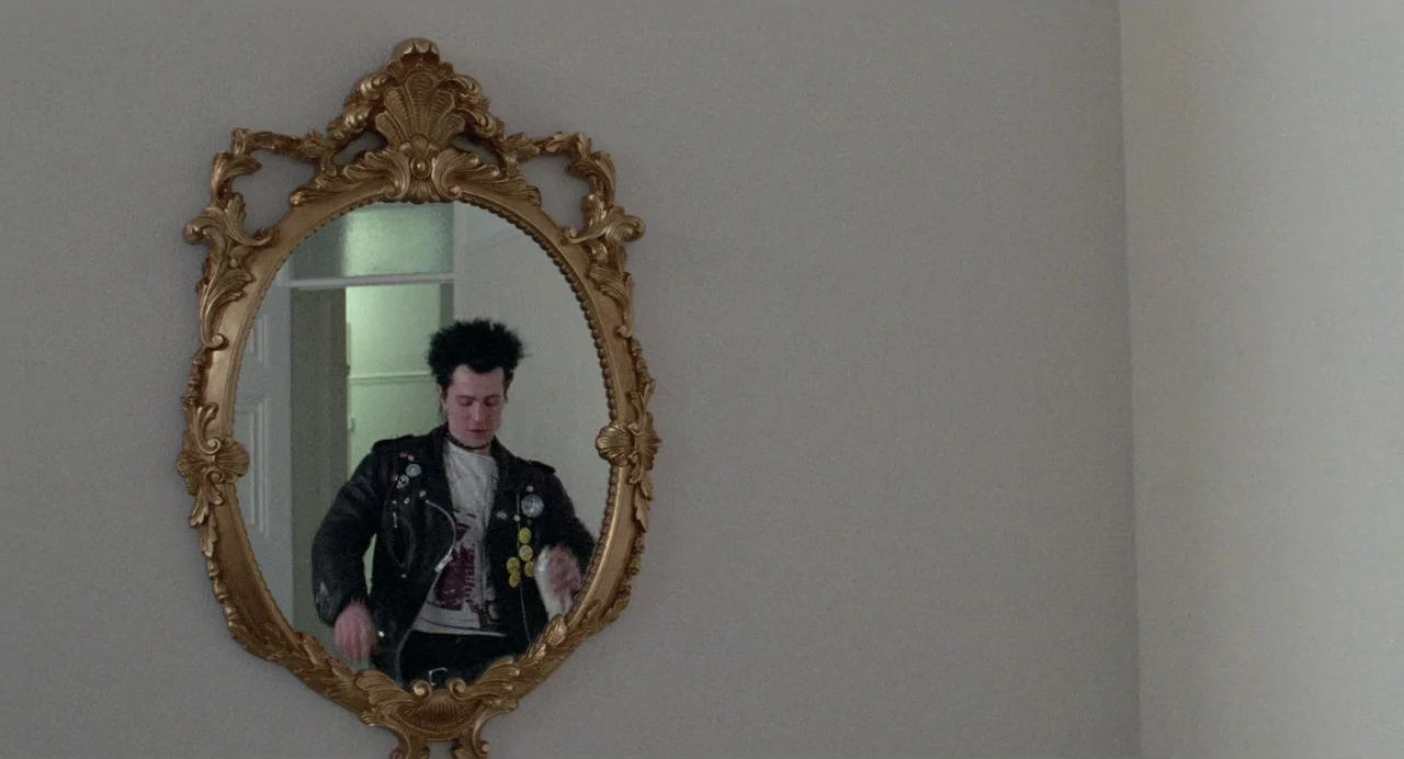 Sid and Nancy (1986) movie still: medium — A man with spiked hair is visible in a large ornate gold-framed wall mirror. The mirror hangs on a plain,…