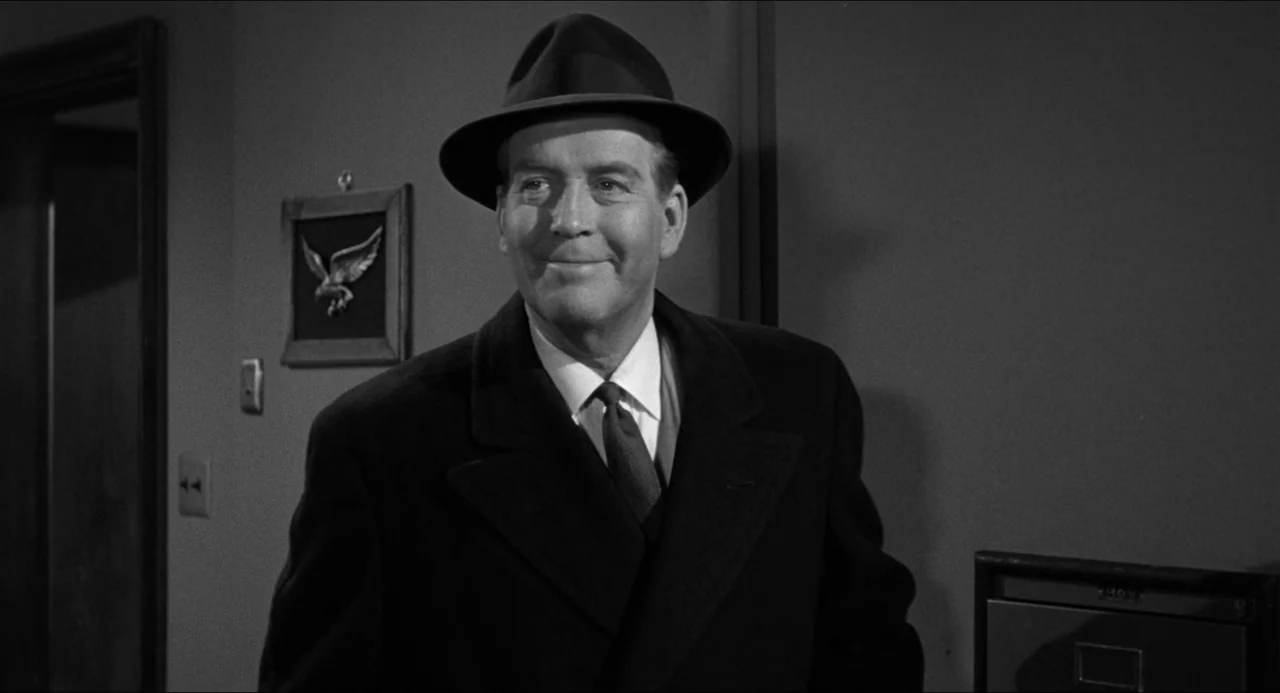 The Fearmakers (1958) movie still: medium — An interior space with a blank wall featuring a small eagle framed picture. A man in a dark hat and coat…