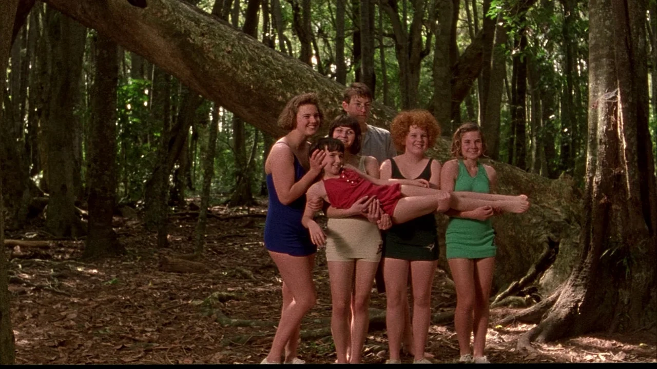 An Angel At My Table (1990) movie still: fullbody — A group of teenage girls stands in a dense, sun-dappled forest clearing. They are posing…