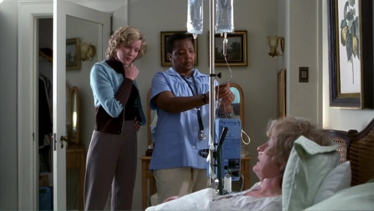 Music from Another Room (1998) movie still: medium — A well-lit room contains a hospital bed and a portable IV stand. A nurse assists a patient in…