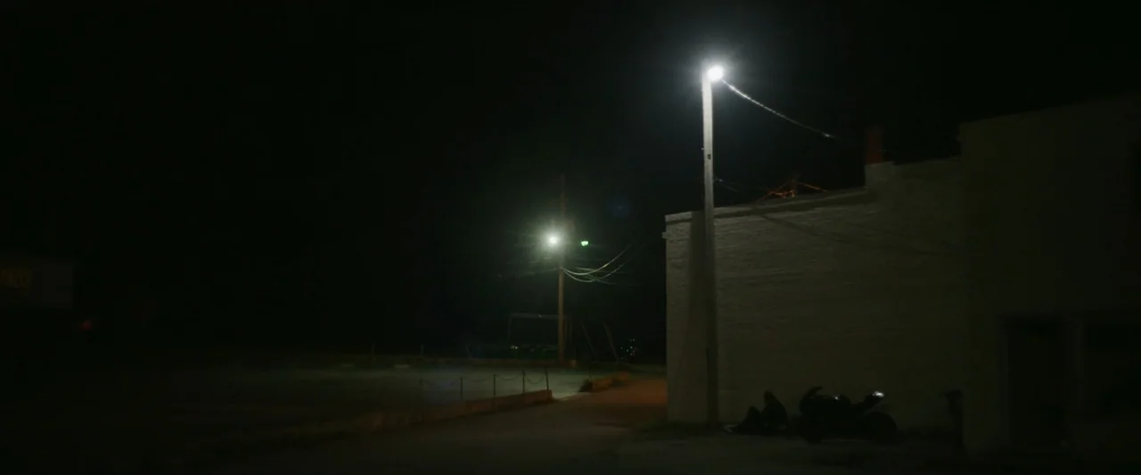 One Fast Move (2024) movie still: wide — An empty parking lot at night sits beside a plain white masonry building wall. A single streetlamp…