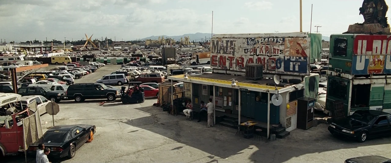 The Hangover (2009) movie still: wide — A dusty auto salvage yard contains hundreds of wrecked vehicles and a weathered office. Three men sit on the…