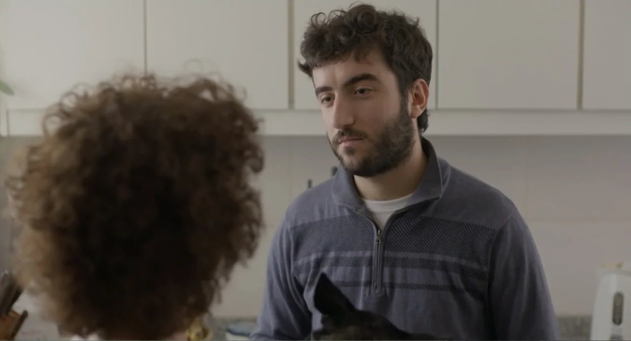 An Odd Turn (2024) movie still: medium — A modern kitchen setting with white cabinetry visible in the background behind the subject. A man in a blue…
