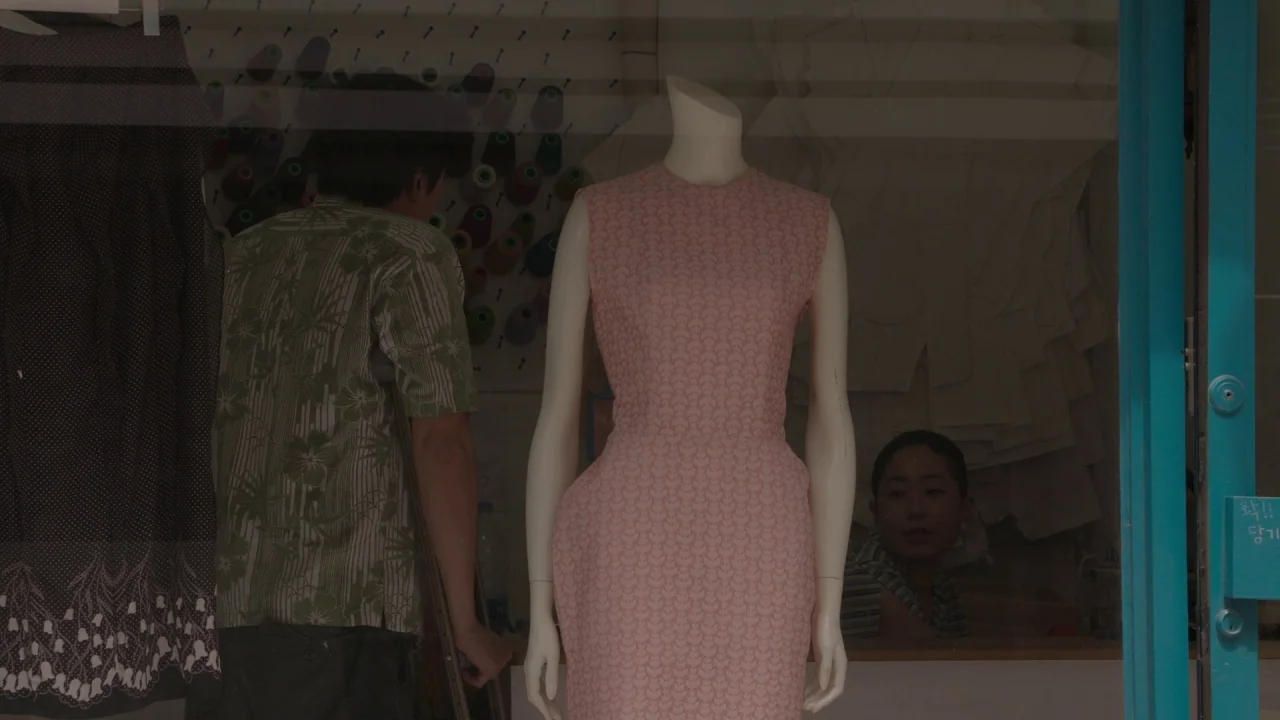 Yourself and Yours (2016) movie still: medium — A tailor shop window displays a headless mannequin wearing a pink patterned dress. A man in a green…