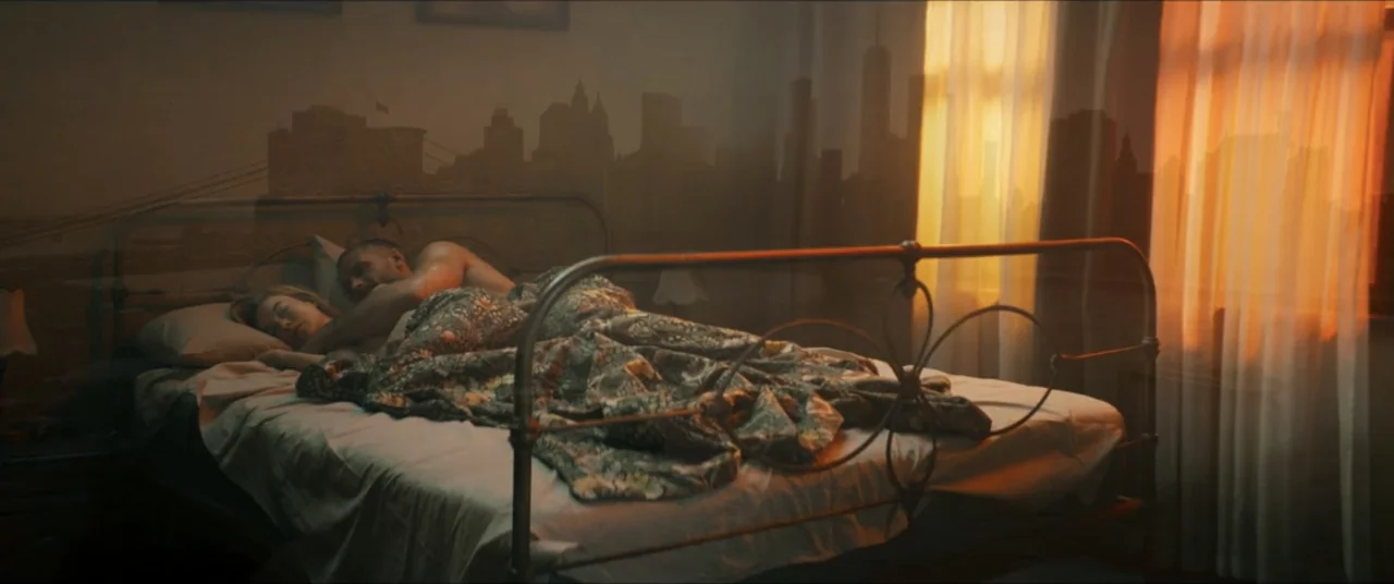 Man with No Past (2025) movie still: medium — A sunlit bedroom features a metal bed frame against a silhouetted city skyline backdrop. A man and a…