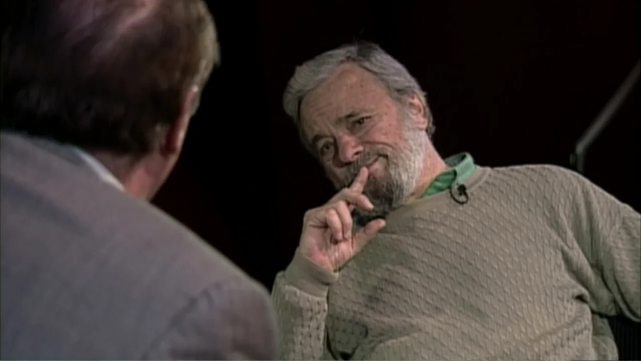 Six by Sondheim (2013) movie still: medium — A man sits on a set during an interview against a dark void background. The man in a tan sweater rests…