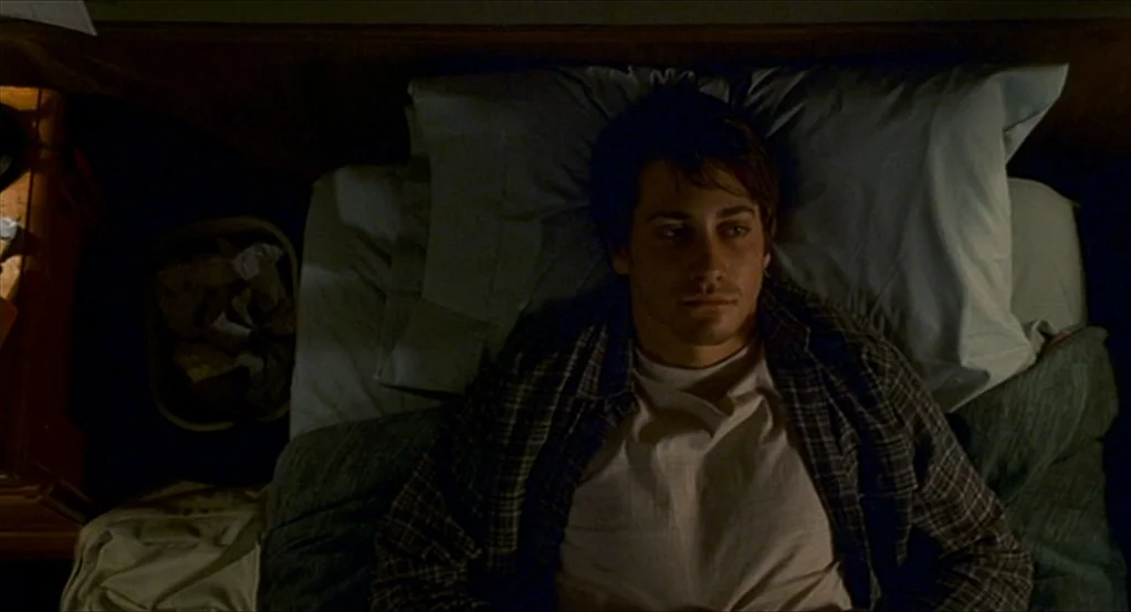 The Good Girl (2002) movie still: medium — A dark, dimly lit space with a person resting on a simple mattress. The individual is positioned directly…