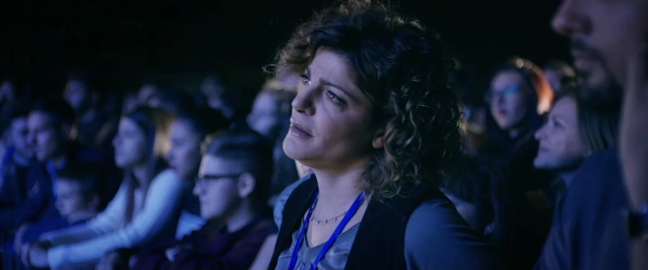 Taranta On the Road (2017) movie still: medium — An auditorium filled with blurred attendees sitting in rows during a dark presentation. A woman in…