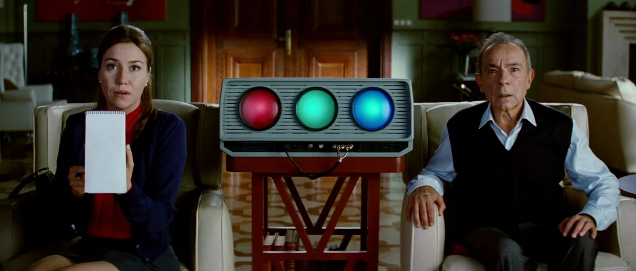 Broken Embraces (2009) movie still: medium — Two individuals sit in leather chairs facing forward in a dark, paneled room. A device with three…