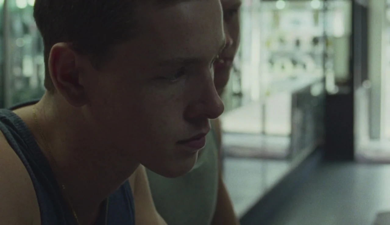Beach Rats (2017) movie still: closeup — A jewelry store interior with glass display cases reflecting ambient light in the background. Two young men…