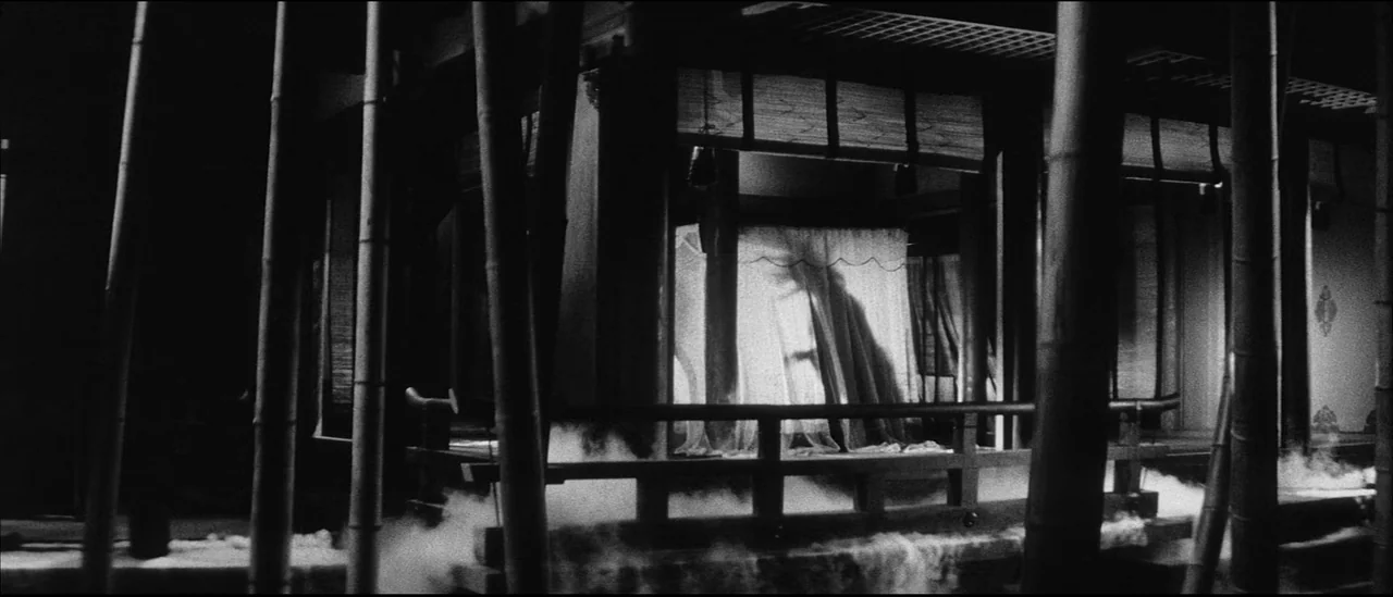 Kuroneko (1968) movie still: wide — A dark wooden structure with traditional sliding doors stands behind a vertical bamboo grove. Low-lying mist…