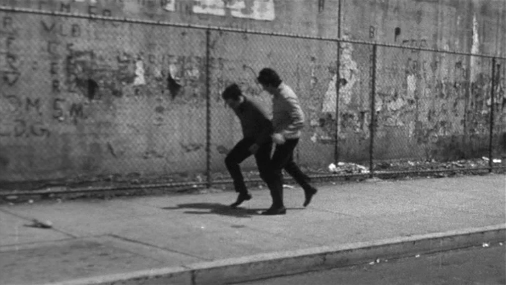 Who’s That Knocking At My Door (1967) movie still: fullbody — A paved sidewalk runs parallel to a tall chain-link fence with graffiti. Two men walk…