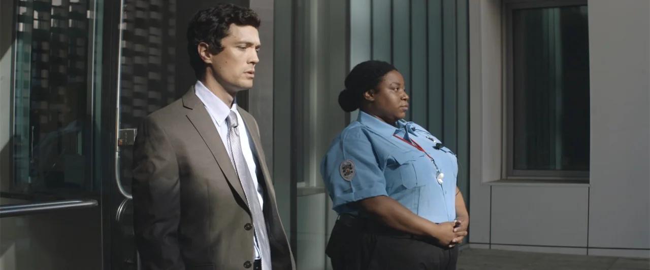 Mennonite's Bite (2023) movie still: medium — A modern building entrance with glass doors and reflective panels features two professionals standing.…