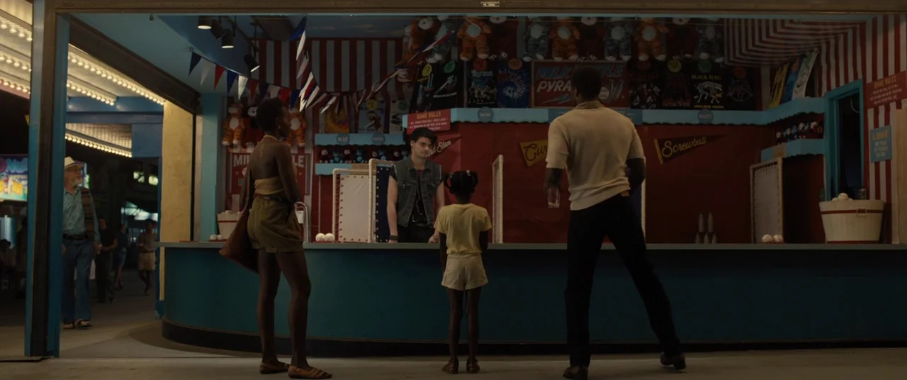 Us (2019) movie still: wide — A nighttime carnival stall with red striped curtains and hanging stuffed animal prizes. A family of three stands…