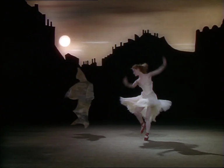 The Red Shoes (1948) movie still: fullbody — A dark, stylized stage set features silhouettes of chimney-topped buildings under a pale moon. A…