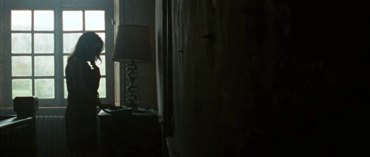 Alléluia (2014) movie still: medium — A dimly lit residential interior features a woman standing by a multi-pane window. She holds a telephone…