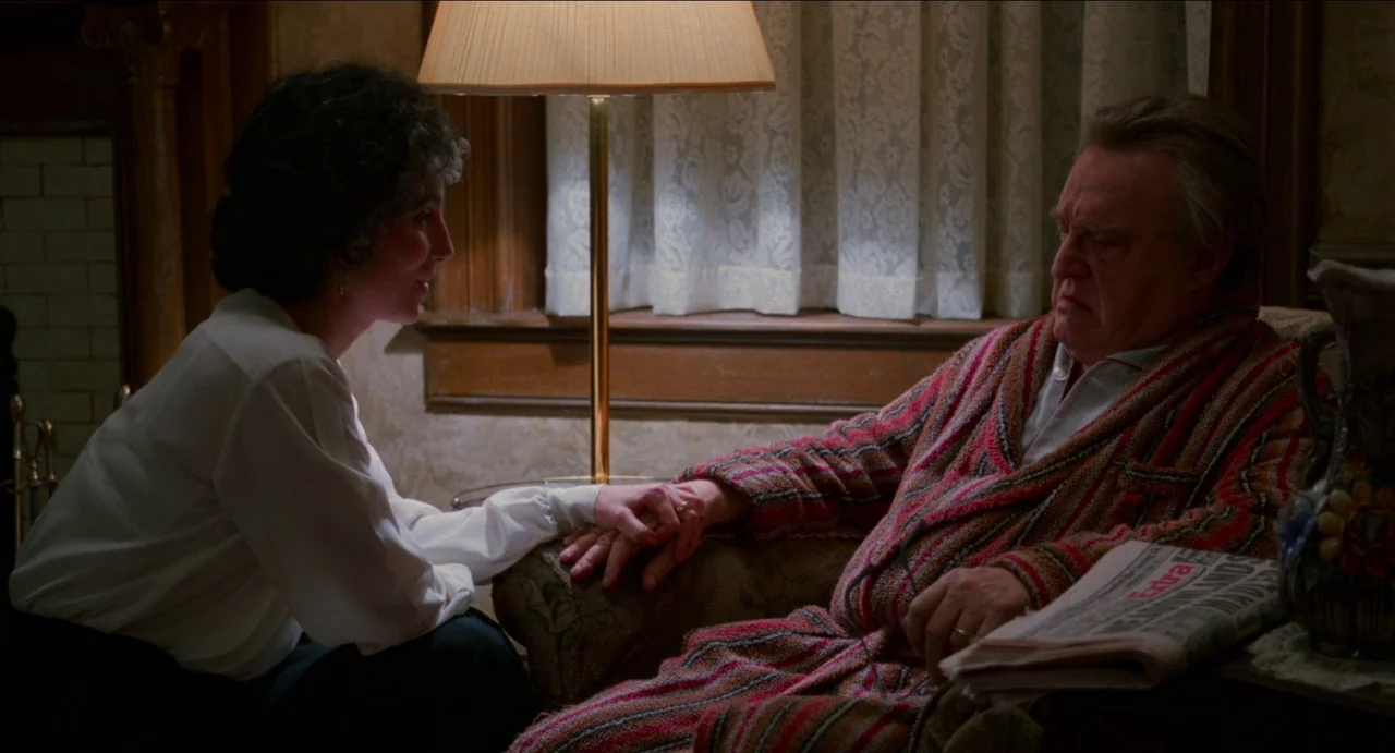 Moonstruck (1987) movie still: medium — An interior space features wood-paneled walls and a lace-covered window behind a lamp. A woman kneels to…