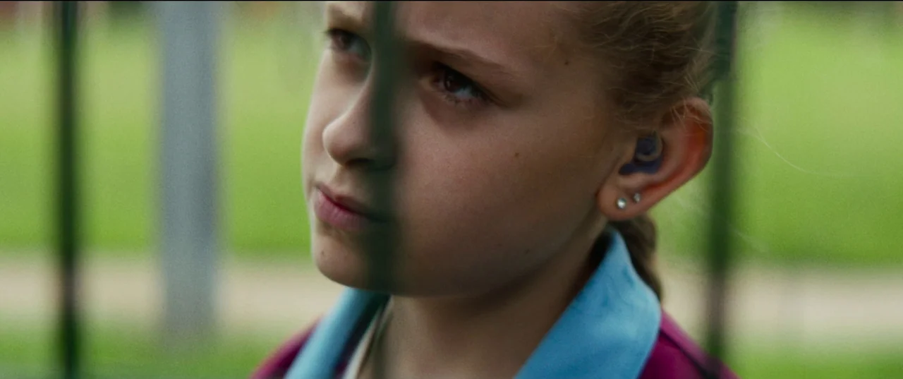 Scrapper (2023) movie still: closeup — A close up view of a girl standing near a wire fence outdoors. The girl looks past a vertical fence wire in…