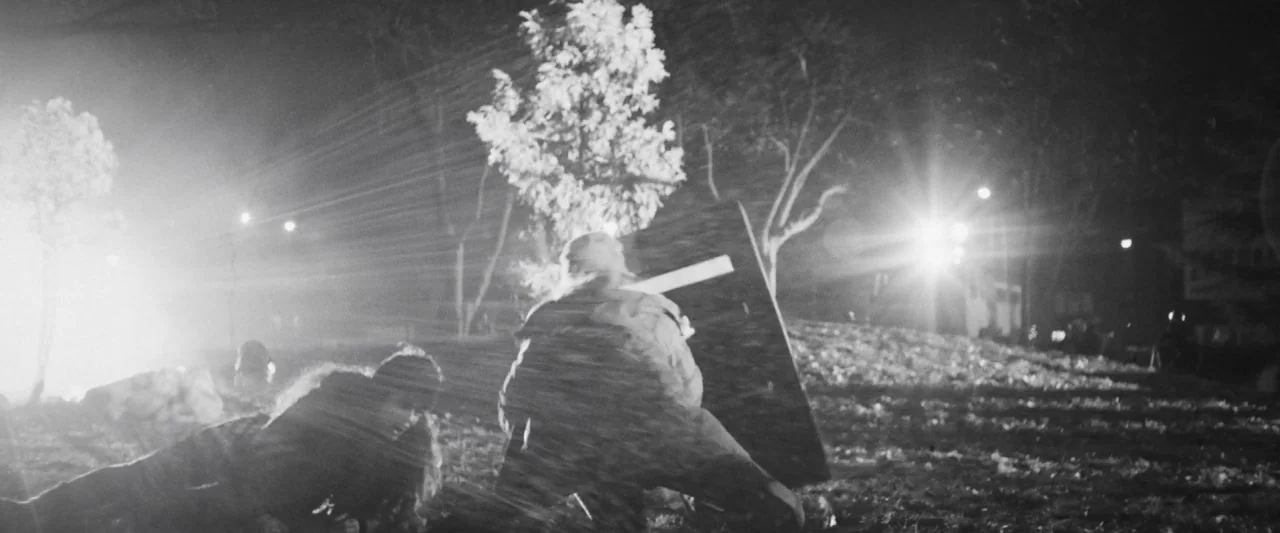 Bushwick (2017) movie still: wide — A dark, grassy park area illuminated by bright, lens-flaring streetlights at night. Two people crouch behind a…