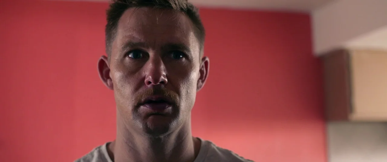 Blindfire (2020) movie still: closeup — A man stands in front of a solid red wall indoors. He stares forward with an expressionless face and his…