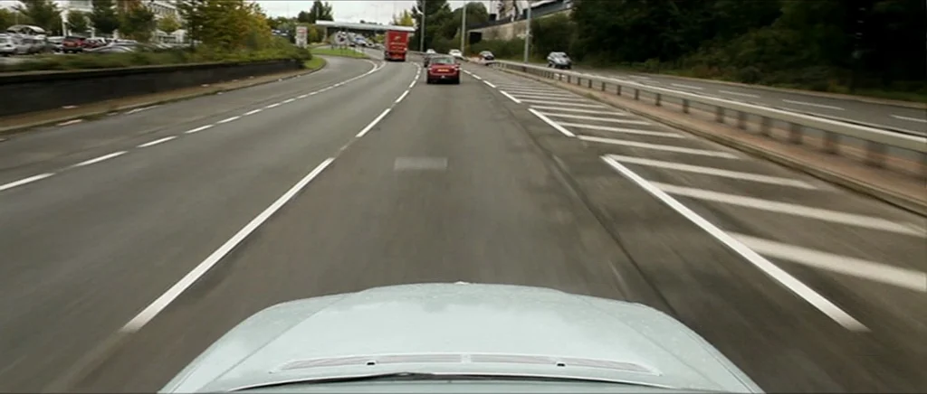 Kill List movie still: wide — A paved multi-lane highway stretches into the distance under an overcast sky. Several cars and a red truck travel…