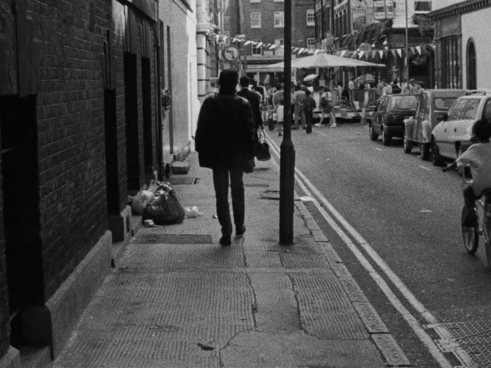 Following (1998) movie still: fullbody — A narrow urban street lined with brick buildings and parked vehicles during the day. A person walks down…
