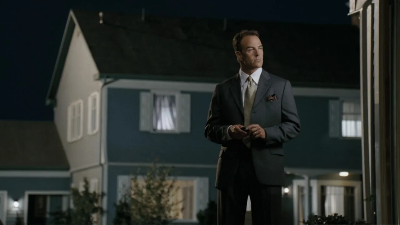 Fun with Dick and Jane (2005) movie still: medium — A two-story blue suburban house at night with illuminated windows sits in the background. A man…