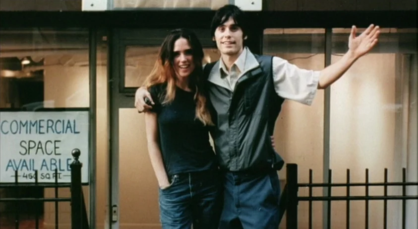 Requiem For A Dream (2000) movie still: medium — A pair stands in front of a glass storefront with a rental sign visible. The young man has his arm…