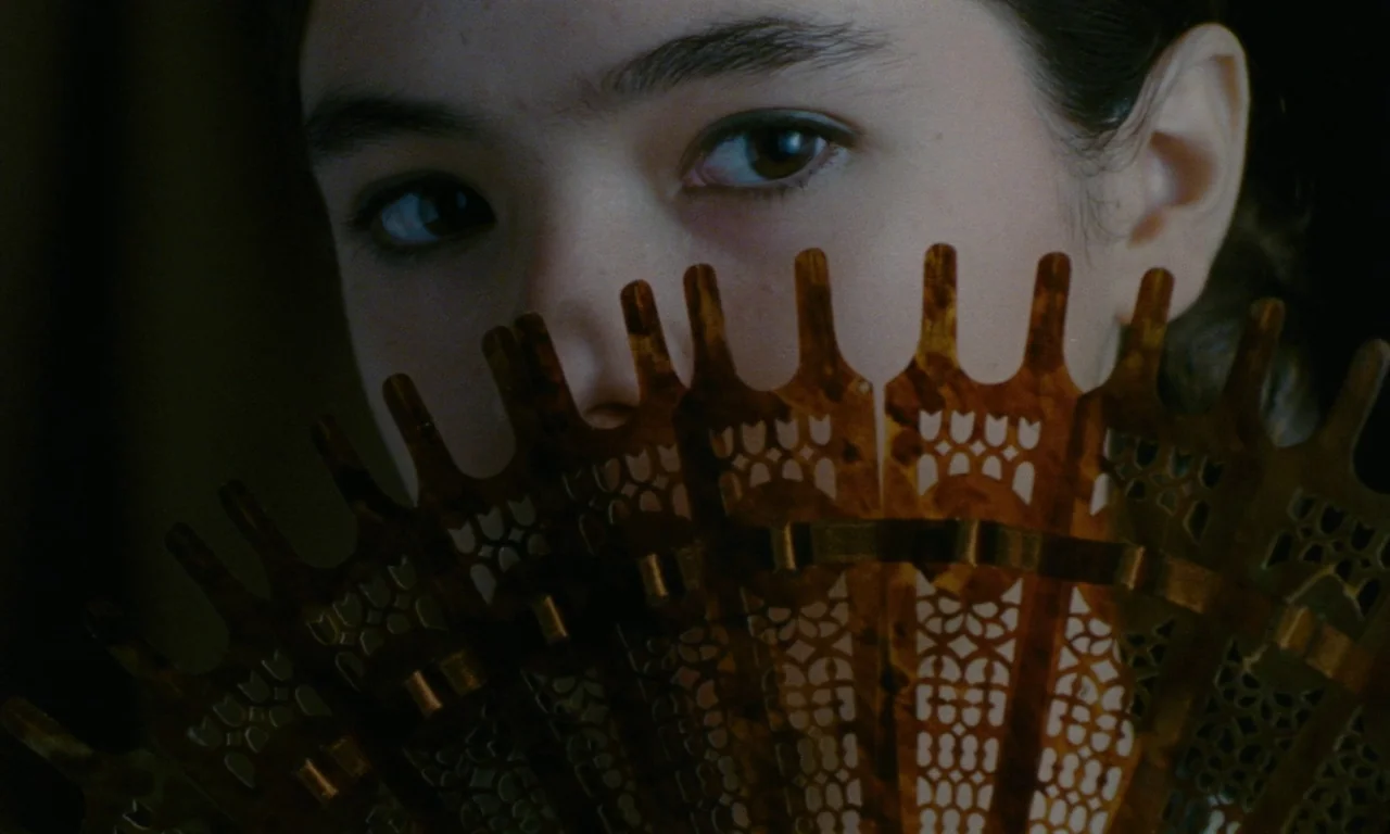 Close Your Eyes (2023) movie still: closeup — A dimly lit indoor space with focus on a woman's eyes. A patterned brown fan obscures the lower half…