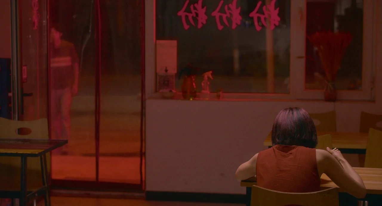 White River (2023) movie still: medium — A quiet restaurant at night with red neon signage visible on the glass window. A woman sits at a table…