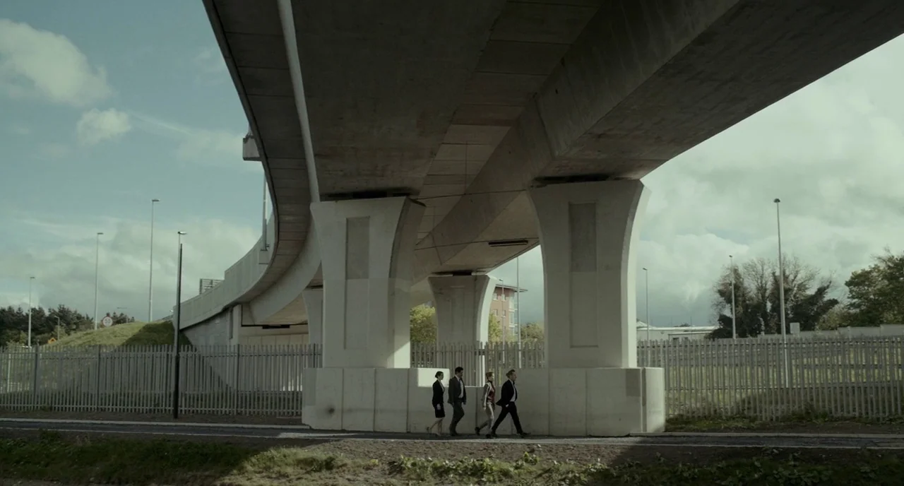 The Lobster (2015) movie still: wide — Four people in business suits walk along a path beneath a large concrete bridge. The site is bounded by tall…
