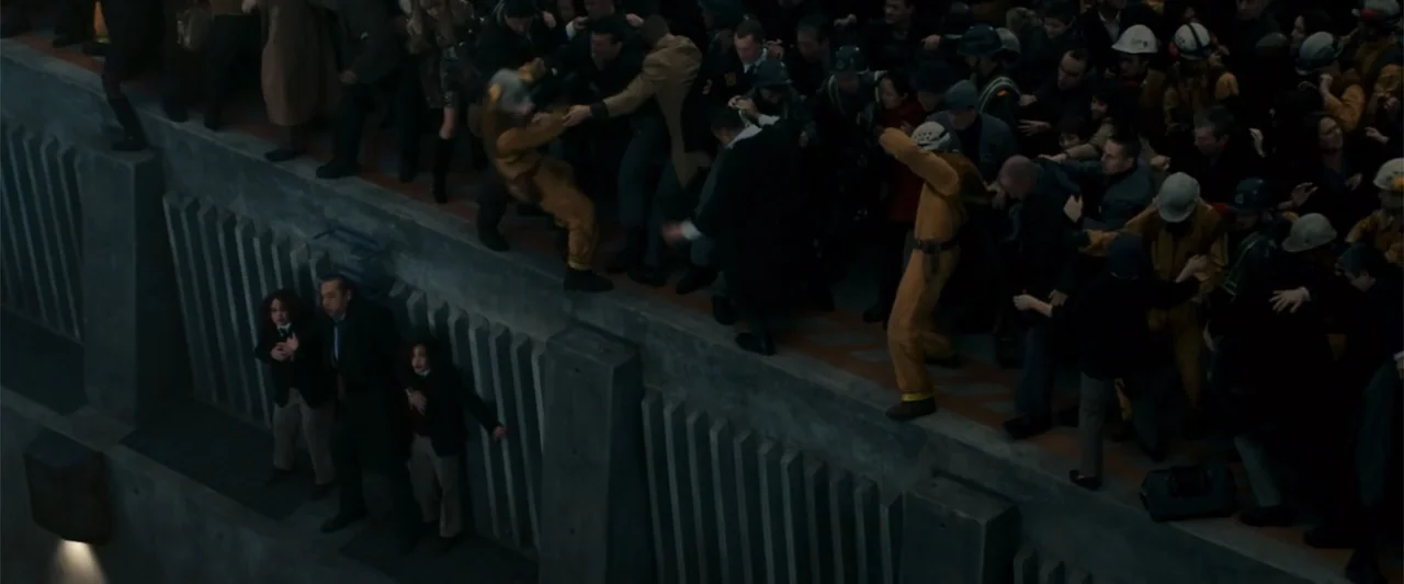 2012 (2009) movie still: wide — A crowded concrete walkway spans the side of a large, brutalist structure at night. People in varying work uniforms…