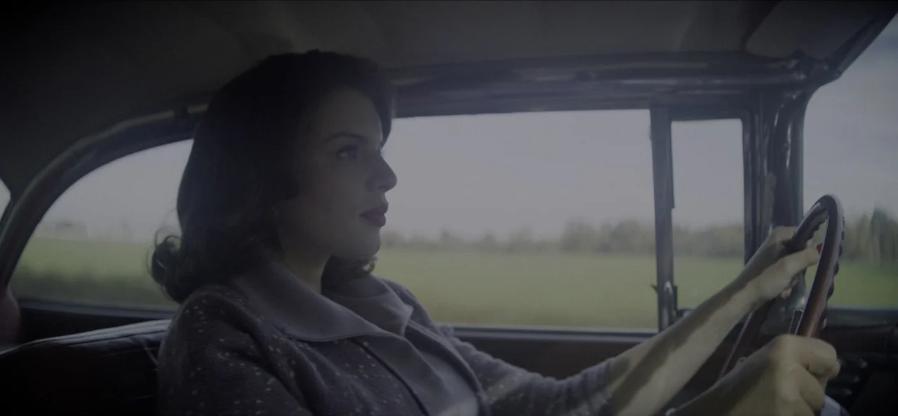 No Sudden Move (2021) movie still: medium — A woman drives a vintage car through a rural landscape during daylight. She maintains her focus on the…