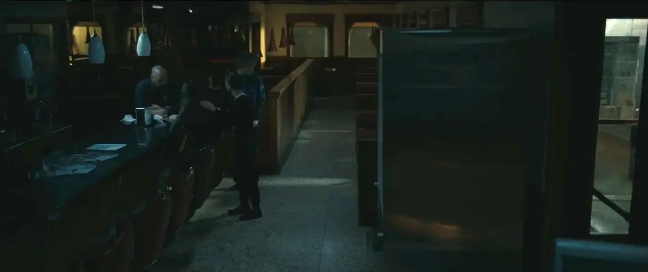 Guns Up (2025) movie still: wide — A dimly lit, near-empty 1950s style diner with tiled floors and vinyl booth seating. Three individuals congregate…