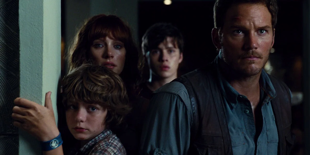 Jurassic World (2015) movie still: medium — A dimly lit indoor corridor with concrete pillars and deep shadows. Four individuals of varying ages…