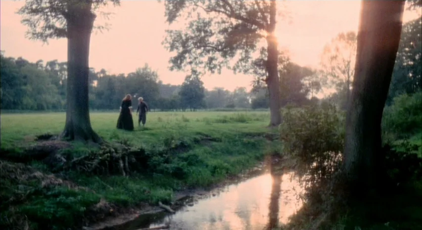 Witchfinder General (1968) movie still: wide — A grassy field features a small winding stream under large mature trees at sunset. Two figures stand…