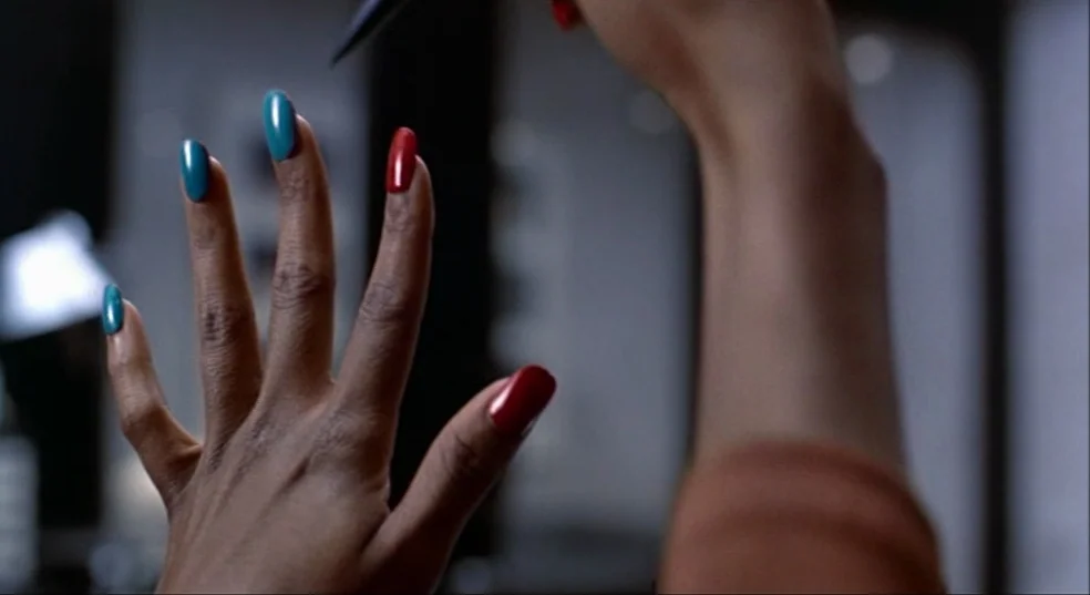 Total Recall (1990) movie still: closeup — A close view of a hand raised against a blurred salon background. The fingers display alternating teal…