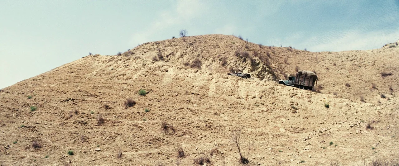 Babylon (2022) movie still: wide — A vast, dry sandy hillside stretches upward under a pale blue sky. Two vintage trucks sit angled on the steep,…