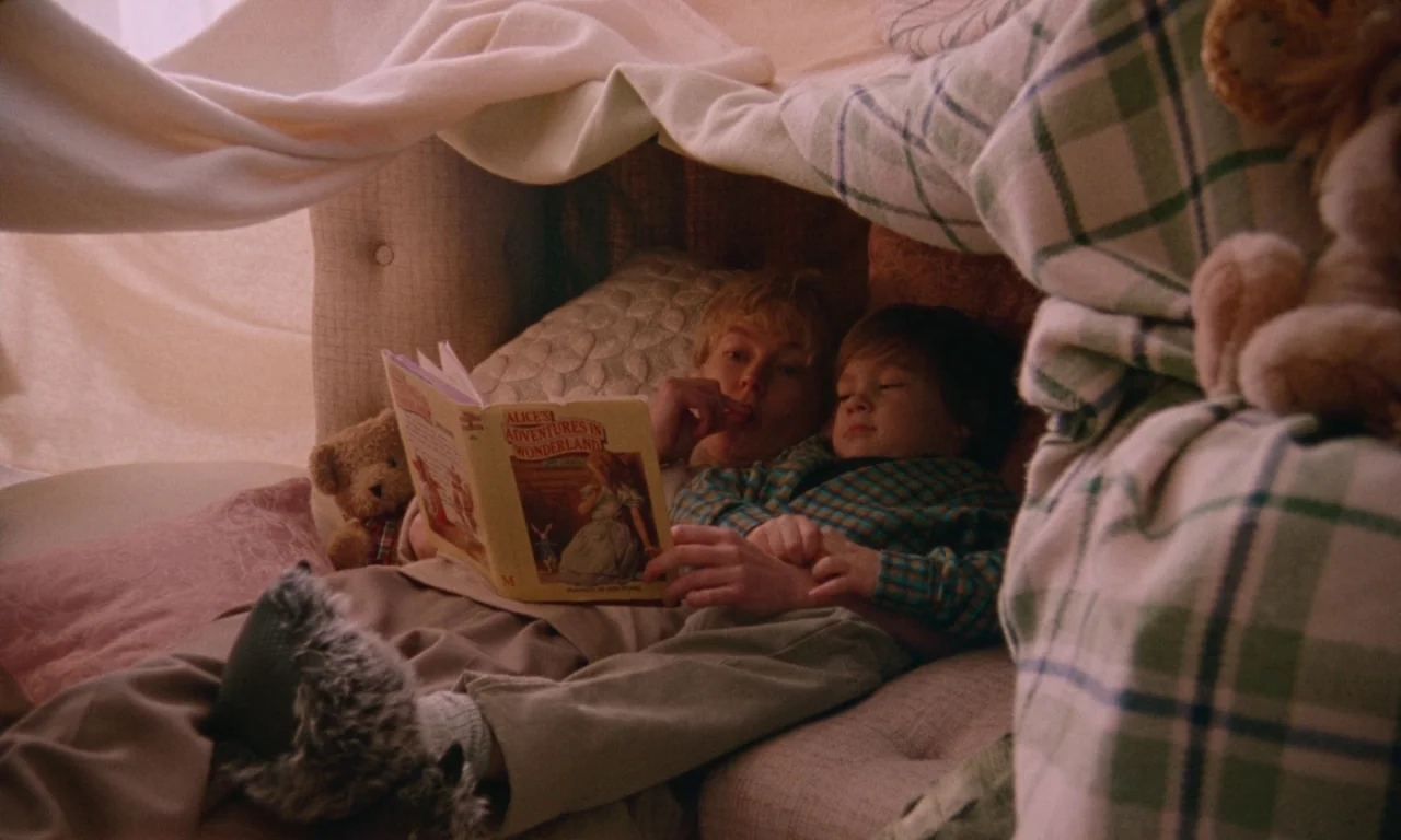 Blue Jean (2022) movie still: medium — A woman and a young boy lie under a makeshift fabric fort indoors. The woman reads an Alice in Wonderland…