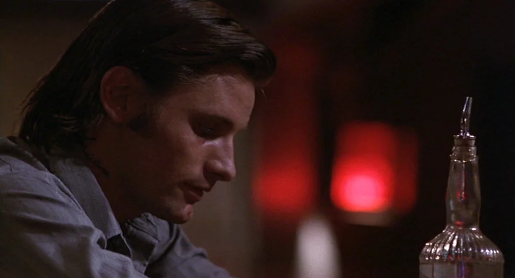 The Indian Runner (1991) movie still: closeup — A dim bar interior with dark wood surfaces and a glowing red neon light. A man sits at a counter…