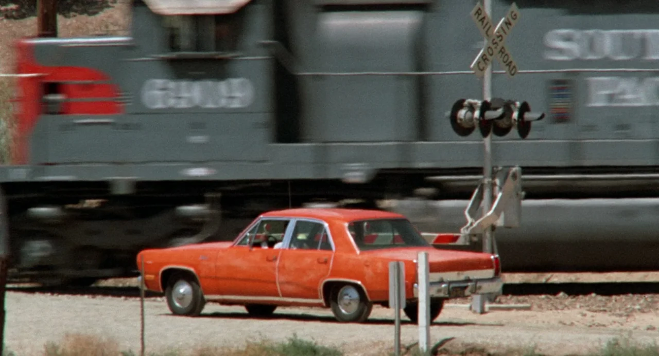 Duel (1971) movie still: wide — A rural level crossing with an active freight train passing in the background. An orange mid-size sedan waits at the…