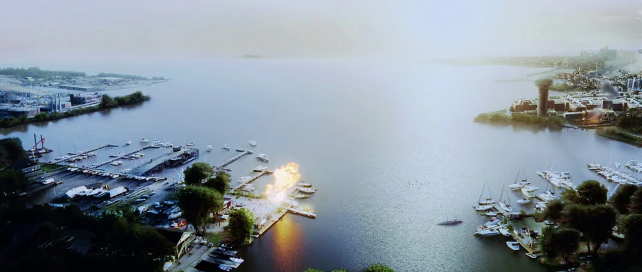 Dawn of the Dead (2004) movie still: establishing — A wide aerial view of a marina on a large body of water. An orange fireball erupts on one of the…