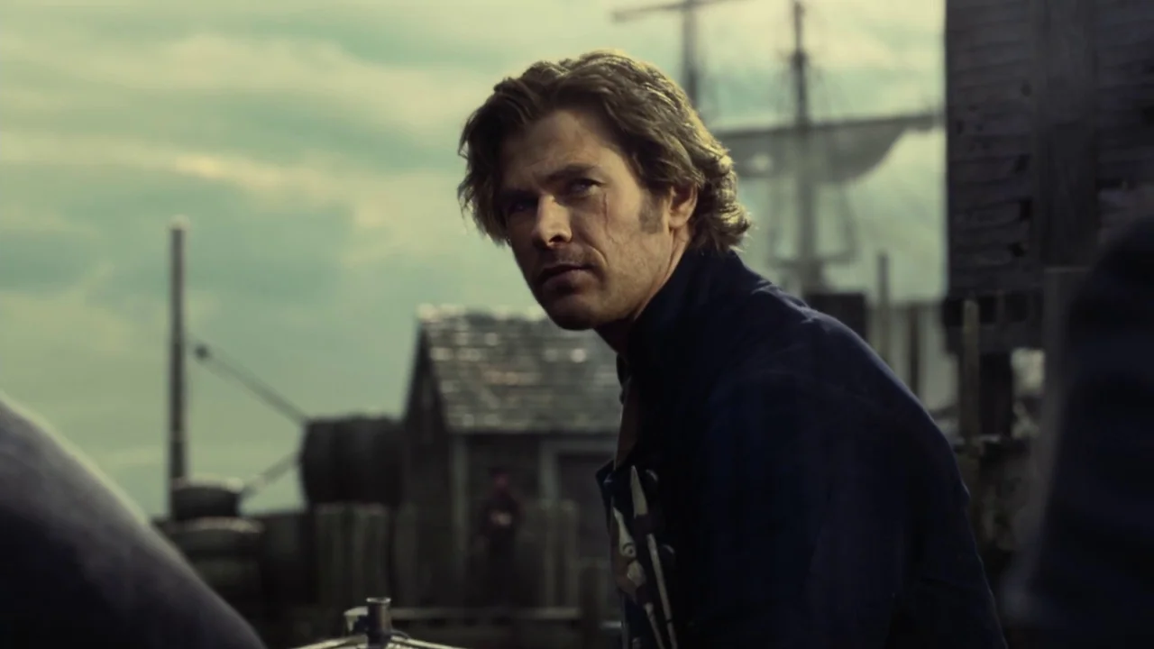 In The Heart of the Sea (2014) movie still: medium — A 19th-century coastal harbor features wooden structures and a ship mast in the background. A…