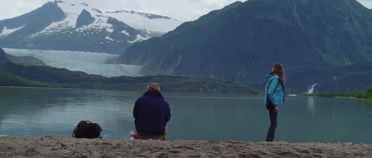 Wildlike (2014) movie still: wide — A wide sandy shore sits before a expansive calm lake and snow-capped mountain range. A man sits on the sand…