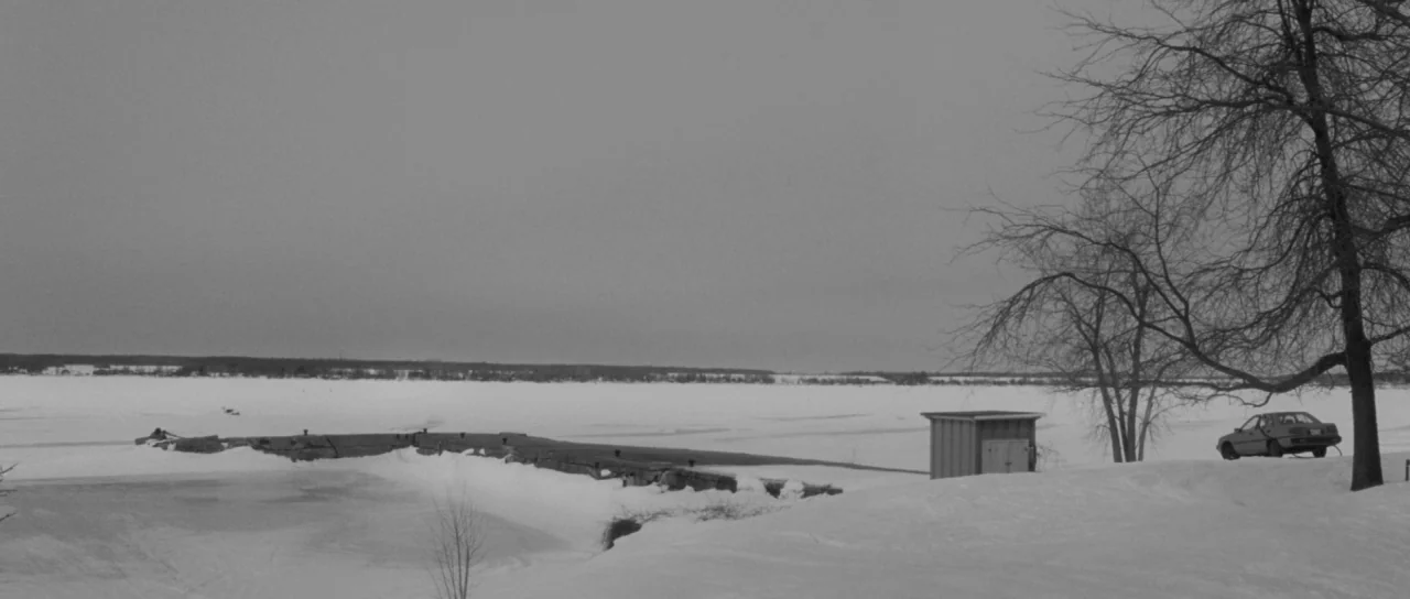 Polytechnique (2009) movie still: establishing — A vast, frozen lake stretches toward the horizon under a heavily clouded grey sky. A lone car is…
