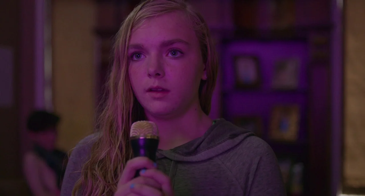 Eighth Grade (2018) movie still: closeup — A dimly lit room with a wooden bookshelf visible in the background. A young woman holds a microphone near…