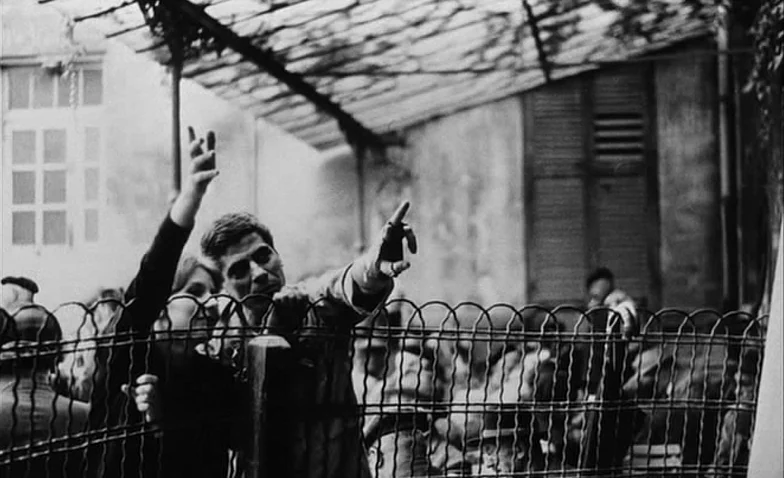 La Jetée (1962) movie still: medium — A man and woman lean over a metal wire fence in an outdoor courtyard. The background features a building with…