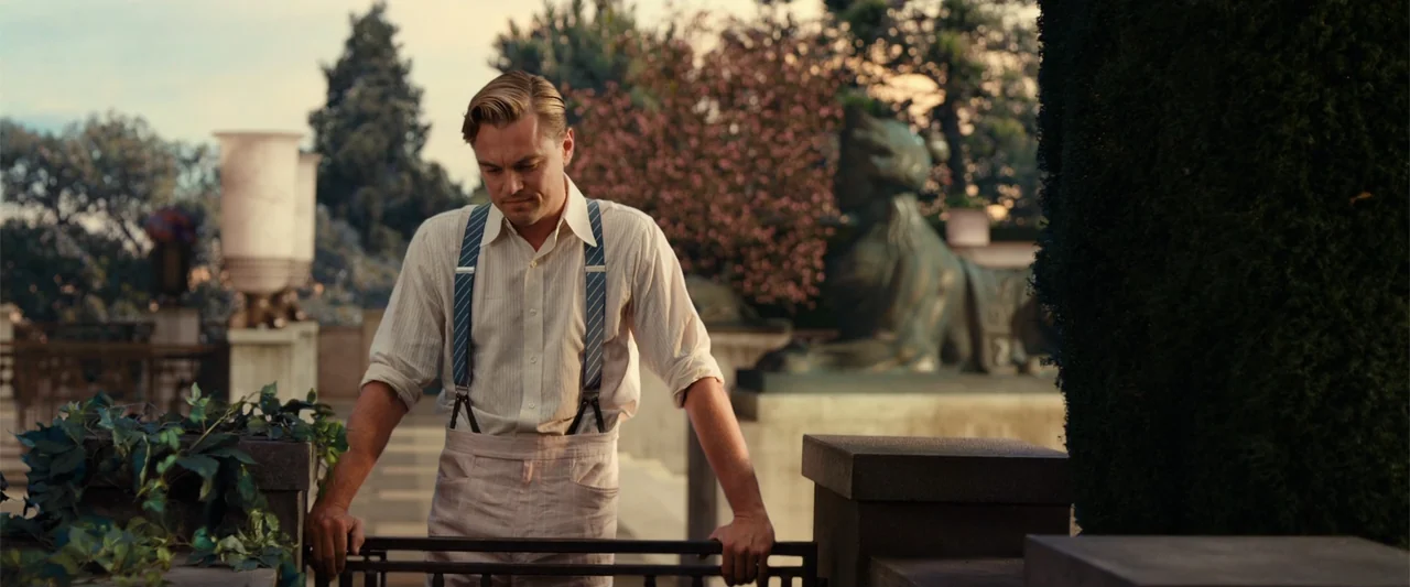 The Great Gatsby (2013) movie still: medium — A man stands on a terrace during sunset with ornamental stone architecture and greenery nearby. The…