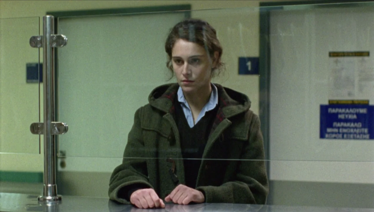 Attenberg (2010) movie still: medium — A sterile building interior with a stainless steel counter and a glass transaction shield. A young woman in…